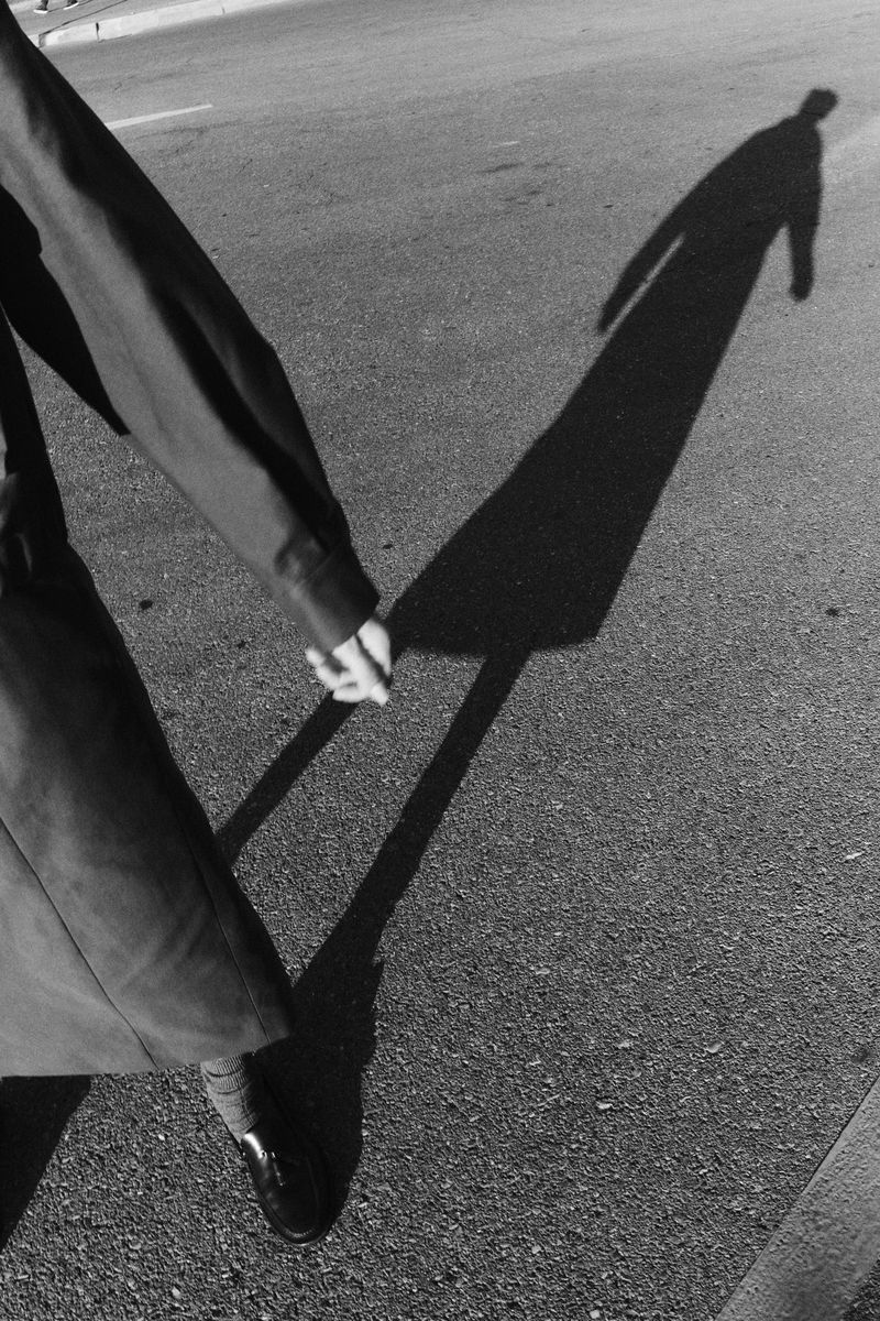 Monochrome photograph captures a person's shadow on pavement, with a foot, hand, and sleeve visible in the foreground, creating a striking composition.