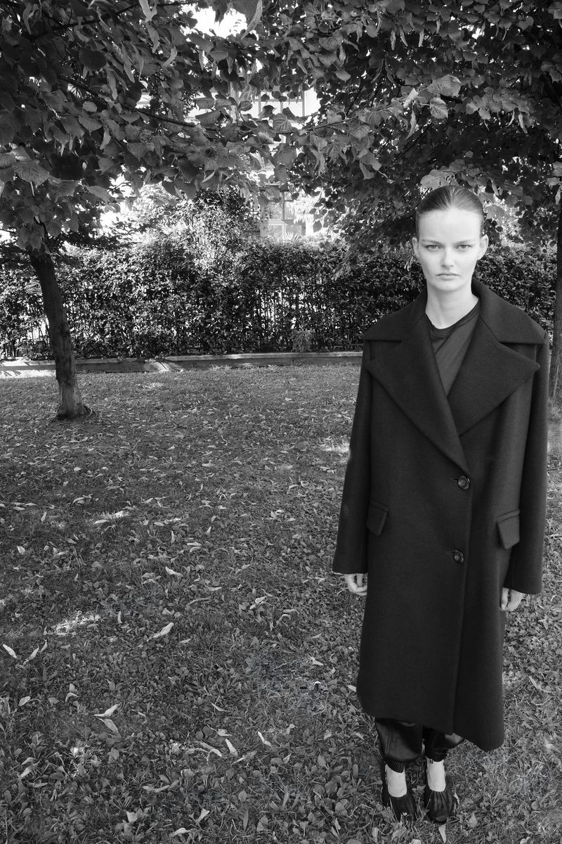 Black and white portrait of a woman in a long coat standing on a grass lawn with trees in the background, in a fashion editorial style.