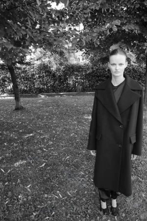 Black and white portrait of a woman in a long coat standing on a grass lawn with trees in the background, in a fashion editorial style.