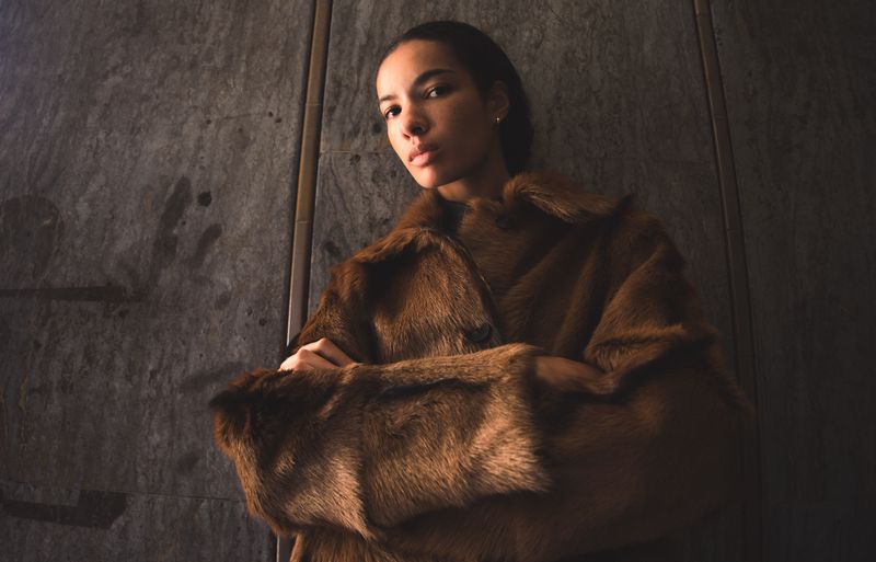 Woman in brown fur coat stands indoors against textured grey wall in medium shot, looking at the camera with neutral expression.