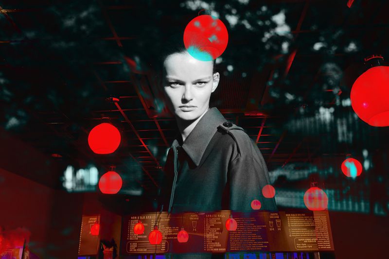 Dramatic fashion shot of an adult woman in a coat with red globe lights in a shadowed interior with a menu board in the foreground.