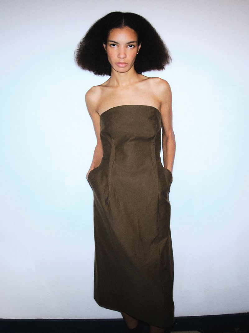 A woman with afro-textured hair poses in a strapless brown dress in a studio against a light blue and white background.