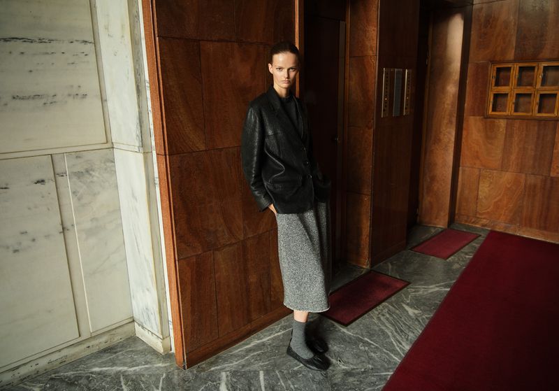 A woman in a black blazer and gray skirt stands indoors with wood-paneled walls and marble floors in a fashion editorial shot.