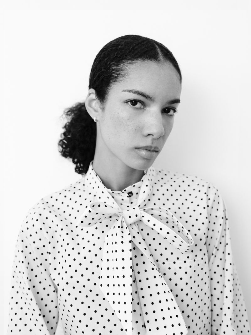 Black and white portrait of a young woman with curly hair wearing a polka-dot blouse with a bow, posing in front of a white background.
