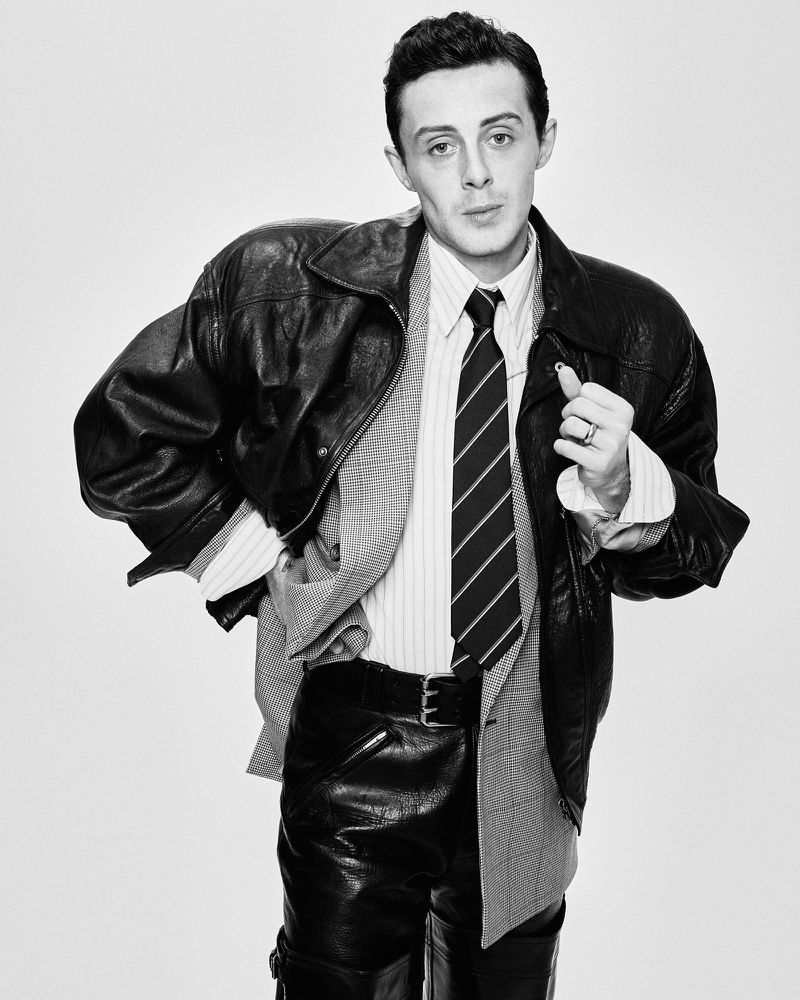 Black and white portrait of a stylish adult male wearing a leather jacket, blazer, tie, and leather pants against a neutral backdrop.