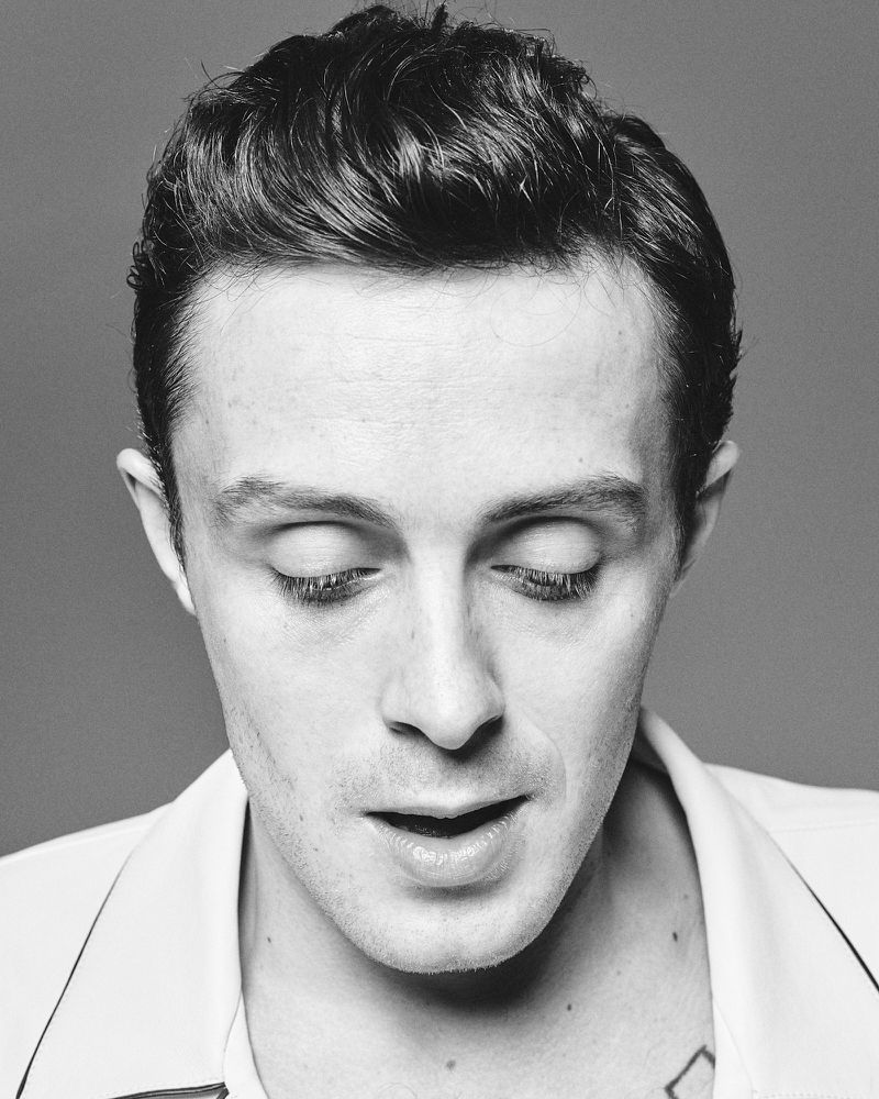 Black and white portrait of an adult male with dark hair, wearing a light colored jacket, and looking down with a neutral facial expression.