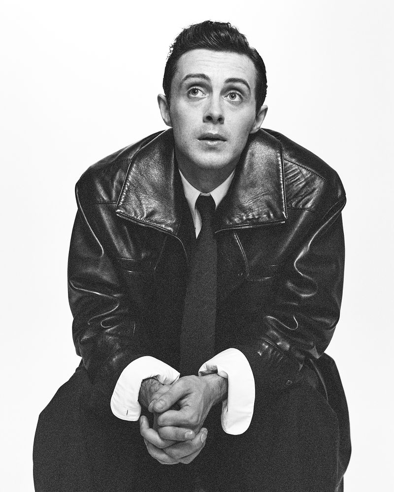 Black and white portrait of adult male wearing a dark jacket and tie, seated against a white background, looking up and to the side.