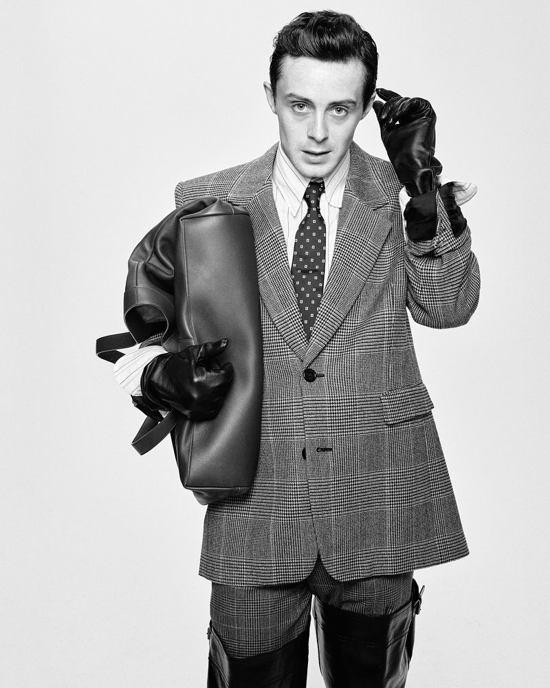 A stylish adult male in a patterned suit holds a leather bag, accented by black gloves and boots, against a clean white background.
