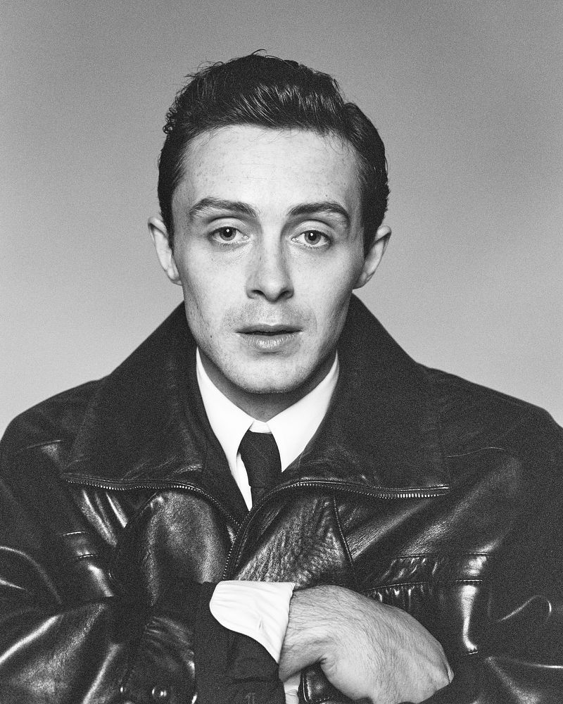 Portrait of model Sonny Hall wearing a dark leather jacket, white shirt, and tie against a grey backdrop.