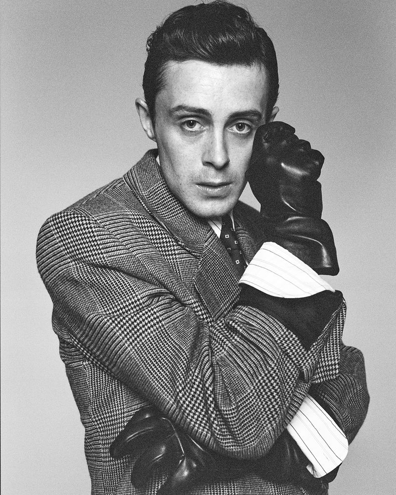 Monochrome portrait of a man in a patterned jacket and leather gloves, looking directly at the camera against a neutral background.