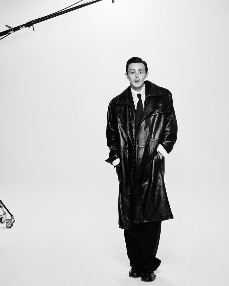 A portrait of a man in a leather coat in a photo studio, with a fair complexion, wearing a dark suit, tie, and black dress shoes.