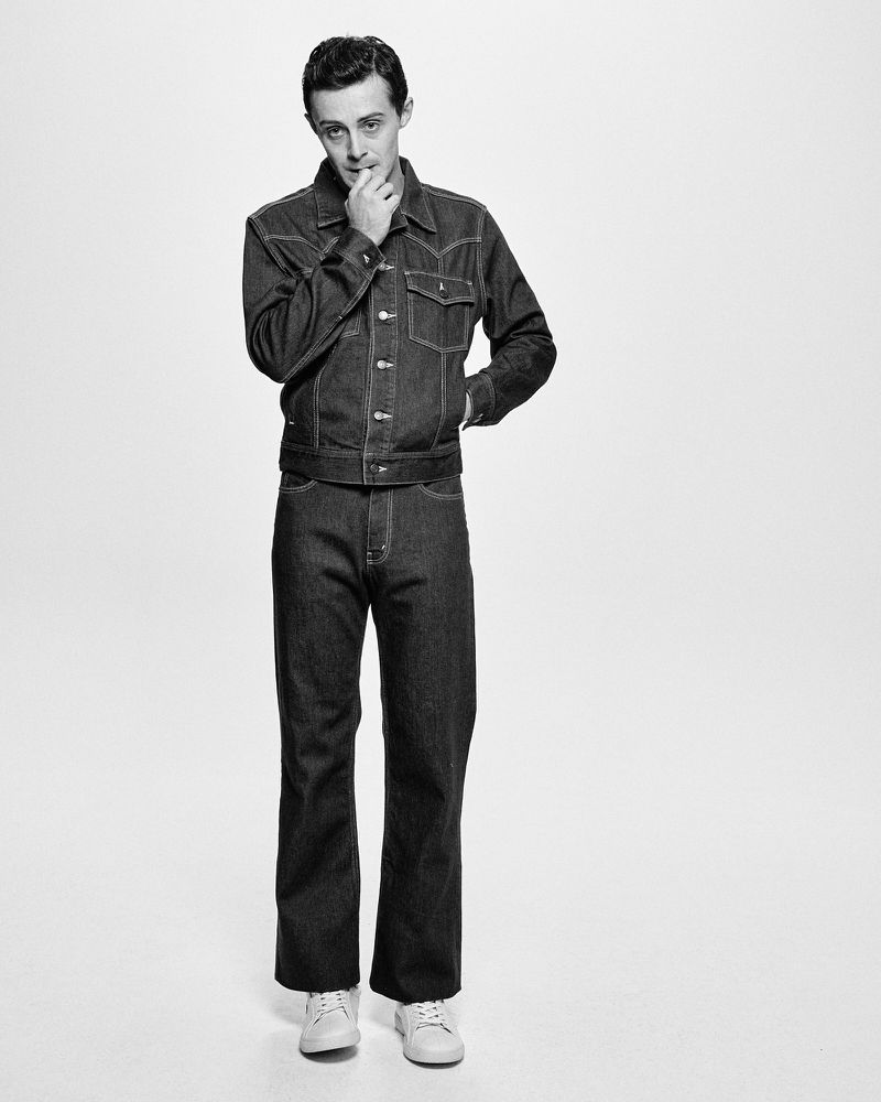 Monochrome portrait of a man in a denim jacket and jeans, standing against a white backdrop in a full shot, with a hand to his mouth.