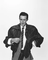Black and white portrait of Sonny Hall wearing an oversized leather jacket, striped shirt, and tie against a plain white background.