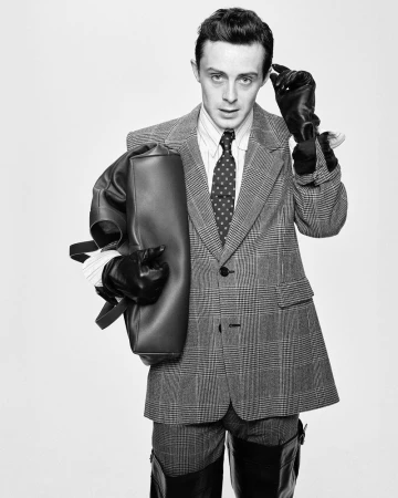 Sonny Hall in a checked suit wearing leather gloves and holding a dark handbag against a plain background