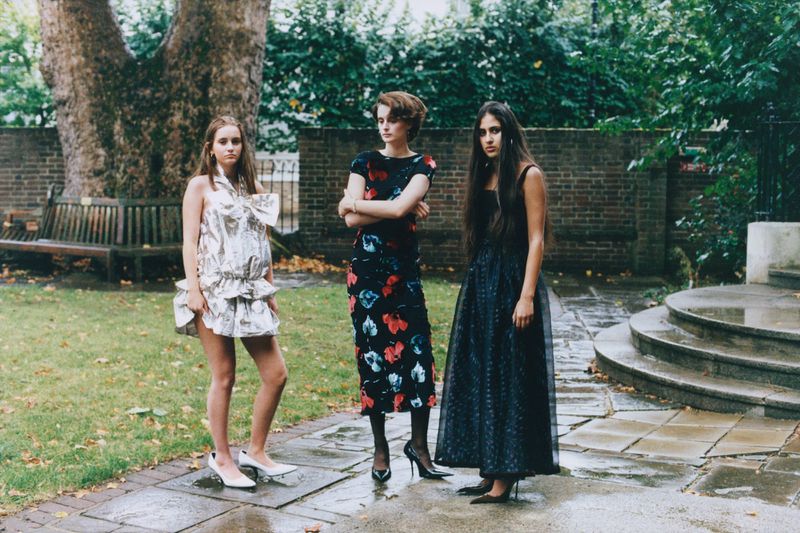 Three women stand in an outdoor garden wearing high fashion dresses, including a metallic dress, a floral dress, and a black dress.