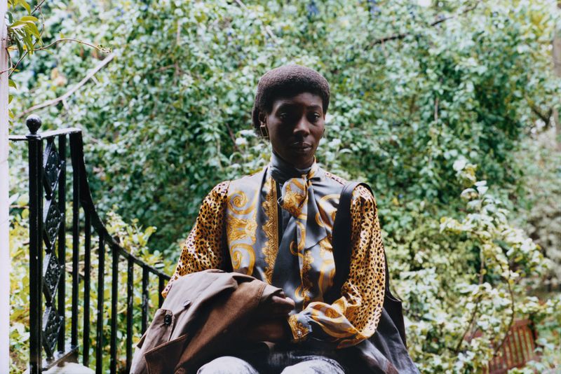 Woman in patterned blouse and light jeans sitting on porch steps surrounded by greenery.  She's holding a brown jacket.