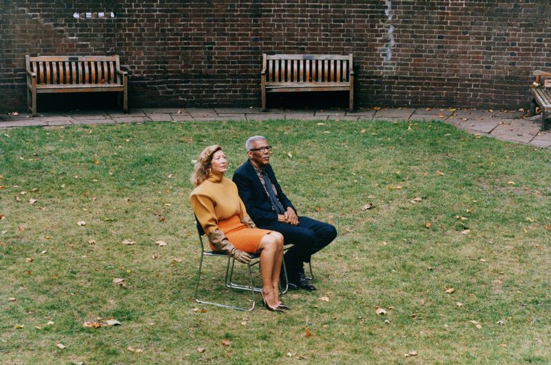 A mature couple sits on metal chairs in a green garden with brick walls and benches, dressed in smart casual attire in daylight.