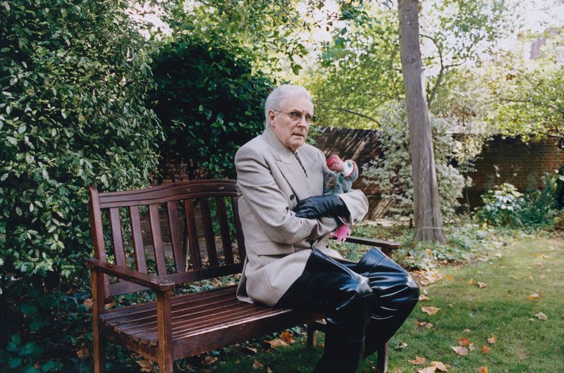 Elderly man in a beige suit sits on a wooden bench in a lush garden, cradling a puppet, looking contemplative amidst the verdant scenery.