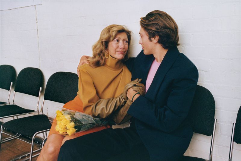 Elegant photo of an older woman in a brown dress and gloves being comforted by a young man in a suit, seated indoors near yellow roses.