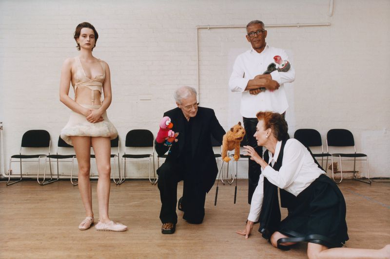Four people pose indoors against a white wall, three holding puppets, including an adult woman kneeling, an elderly man crouching, and a young woman standing.