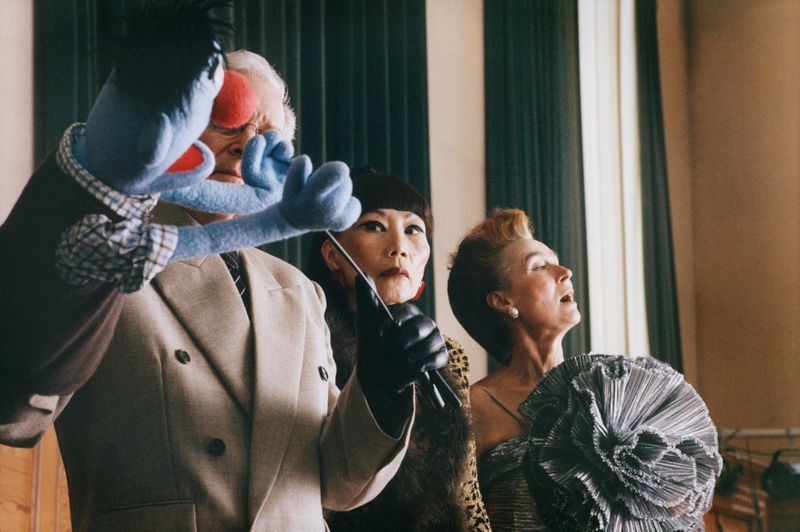 Puppet and people looking up, with an Asian woman in black gloves appearing to conduct them in an indoor setting.