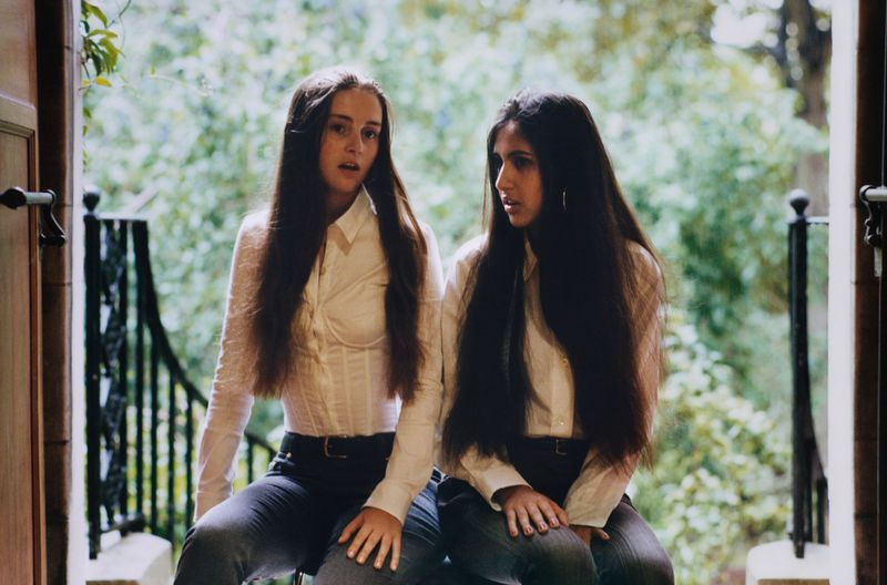 Two young women with long dark hair wearing white button-down shirts and dark trousers pose outdoors in a doorway.