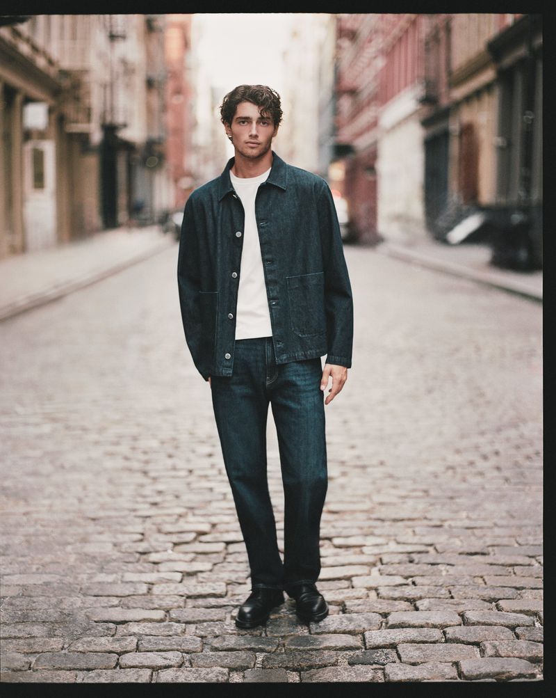Man in denim jacket and jeans stands on cobblestone street, buildings in background, full shot, medium tone, natural light.