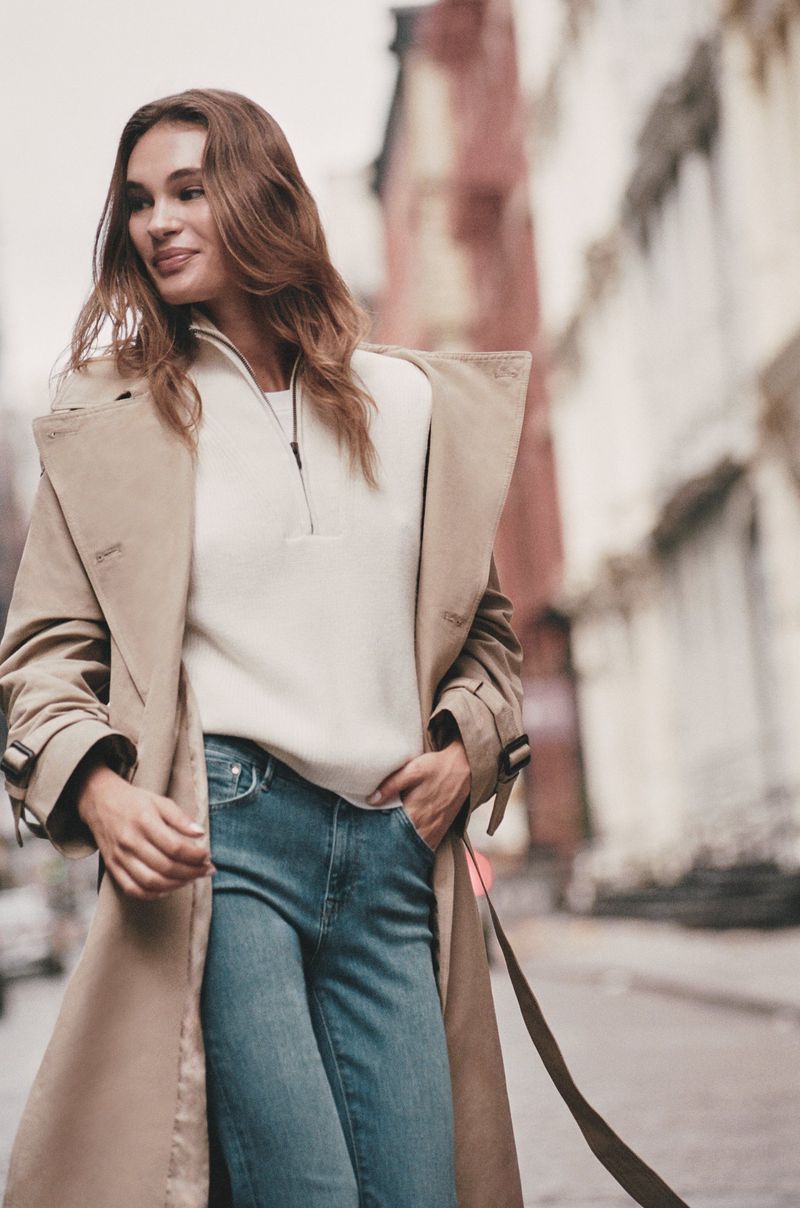 Adult woman in a beige trench coat and blue jeans poses outdoors in a city, smiling, with buildings visible in the background.