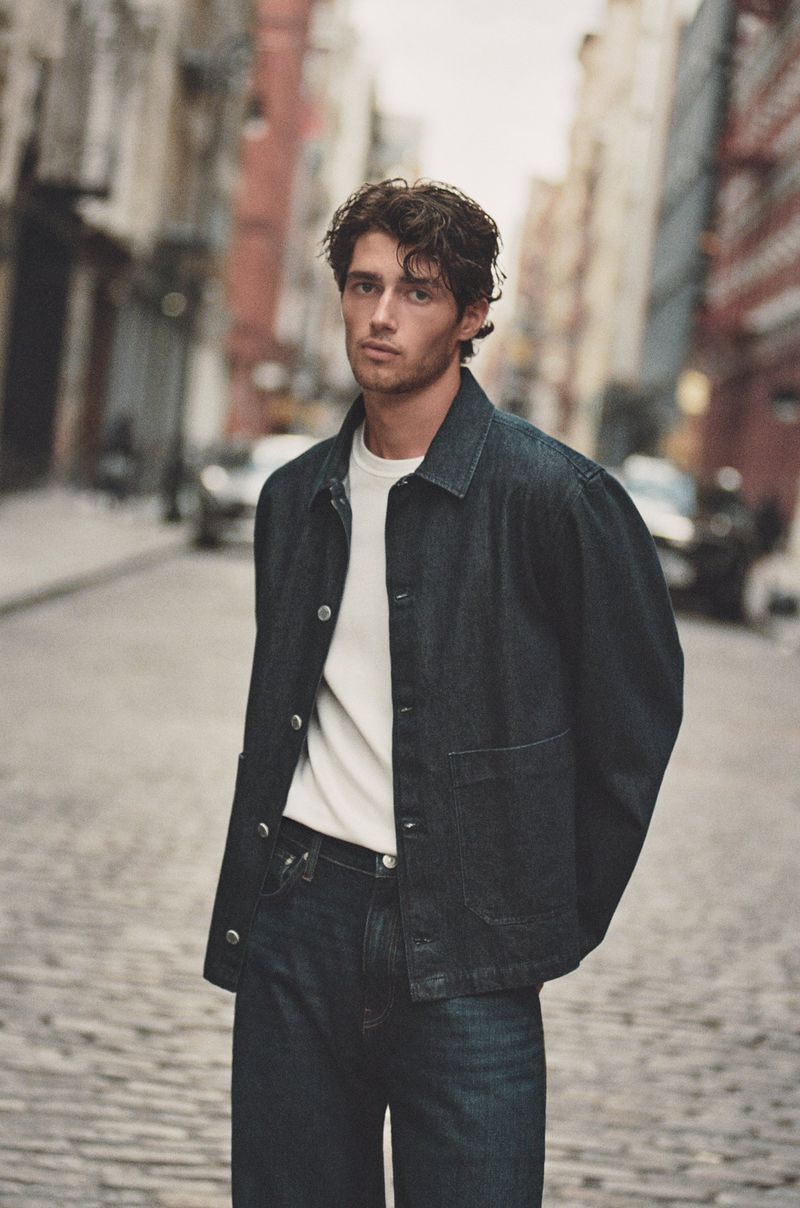 Man in denim jacket and jeans stands on a cobblestone street in a city. Buildings and cars line the street in soft, natural lighting.