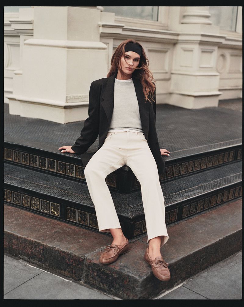A young woman with long brown hair poses confidently on a set of outdoor steps wearing a blazer, jeans, headband, and boat shoes.