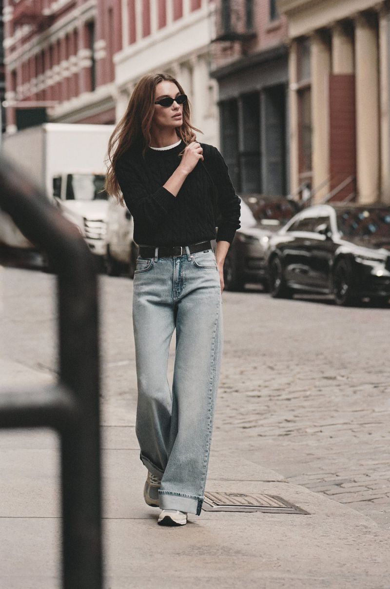 A woman walks on a city street wearing a black sweater, wide-legged jeans, sunglasses, and white sneakers, with cars and buildings visible.