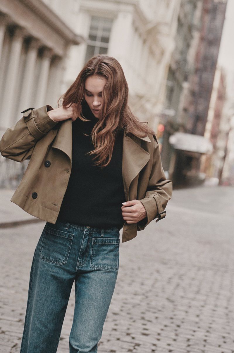 Woman in short trench coat and blue jeans posing in city street with stone buildings on a cloudy day, fashion editorial.
