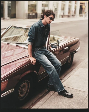 A male model poses on the hood of a burgundy convertible car outdoors, wearing sunglasses, a denim shirt and jeans on a city street.