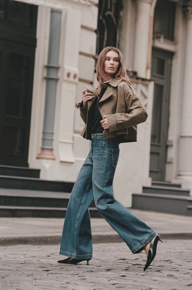 A stylish adult female walks on a city street wearing a beige jacket, wide-leg jeans, and heeled shoes in an outdoor fashion shot.