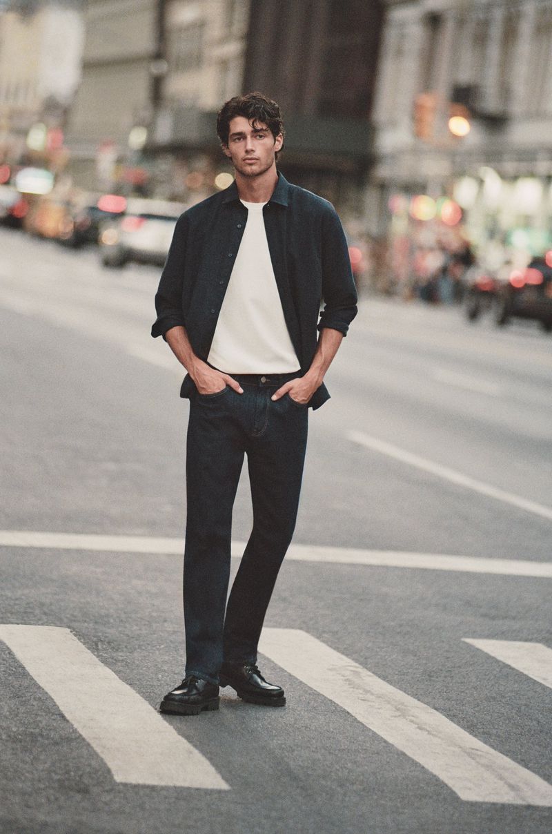 Man poses in dark-wash jeans, white tee, and dark button-down shirt on city street with cars and buildings in blurred background.