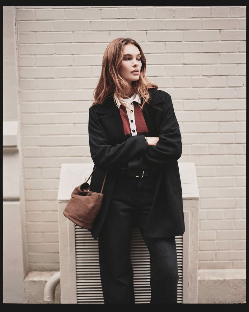 A young adult female in a black coat stands outdoors with a brown purse against a brick wall in an urban fashion portrait.