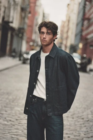 A male model wearing a dark denim jacket and white t-shirt on an urban street for Mavi Fall 2025.