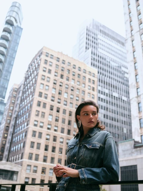 A woman poses wearing a denim jacket in front of modern buildings in an urban setting, creating a fashion-forward and contemporary aesthetic.