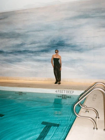 Indoor pool photoshoot: A model in a dark dress and sunglasses poses in front of a mural by the pool in a high-fashion setting.
