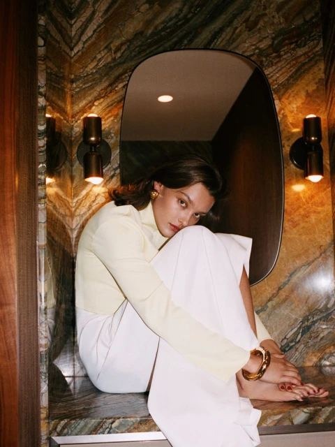 Woman in yellow top and white pants sits on a marble ledge with her feet on a counter in an indoor bathroom setting with wall sconces.