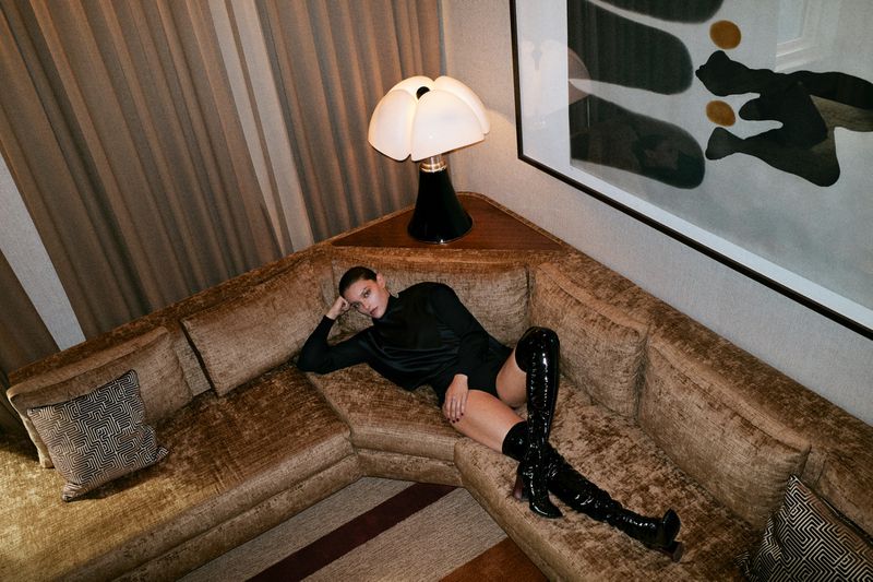Woman in black bodysuit and patent leather thigh-high boots lounges on a gold velvet sectional sofa in an indoor setting.
