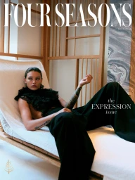 Model lounging on a white chaise lounge wearing a black ruffled top and wide-leg trousers in an interior setting.