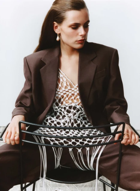 Woman in a brown suit and white crocheted top sits on a black chair against a plain backdrop in a fashion editorial shot.