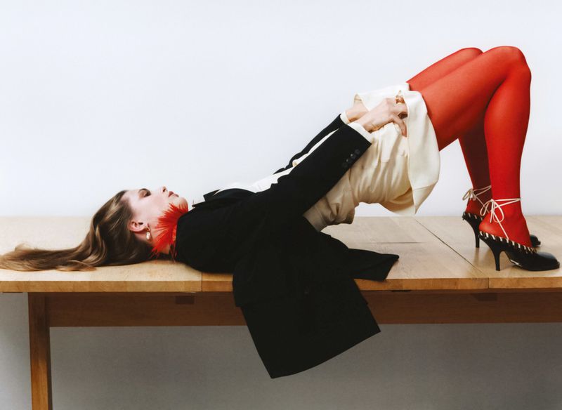 Woman in red tights and heels lies on a wooden table wearing a black and white jacket and a cream-colored dress in a studio setting.