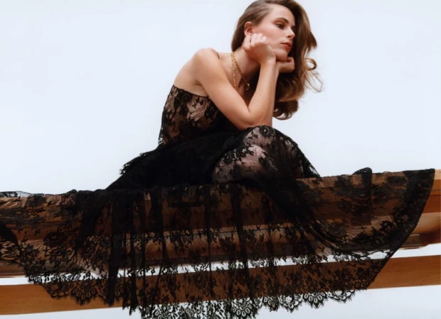Model Lydia-Angel wears a sheer black lace dress while resting on a wooden beam against a pale sky.