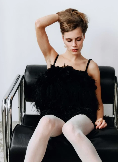 A young adult woman poses while sitting on a black chair against a white wall backdrop, wearing a black top with white tights.