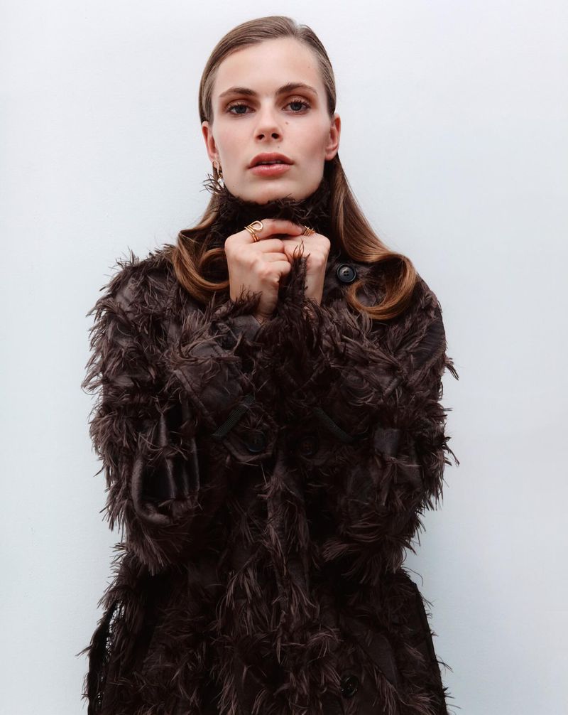Adult woman with light brown hair posing in a dark brown feathery coat against a plain white background in a studio shot.