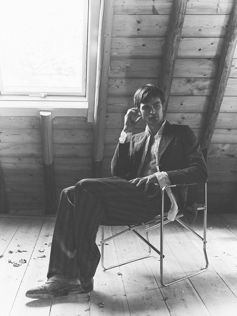 Monochrome portrait of a man in a suit sitting on a folding chair indoors with a window, showcasing a classic and timeless fashion aesthetic.