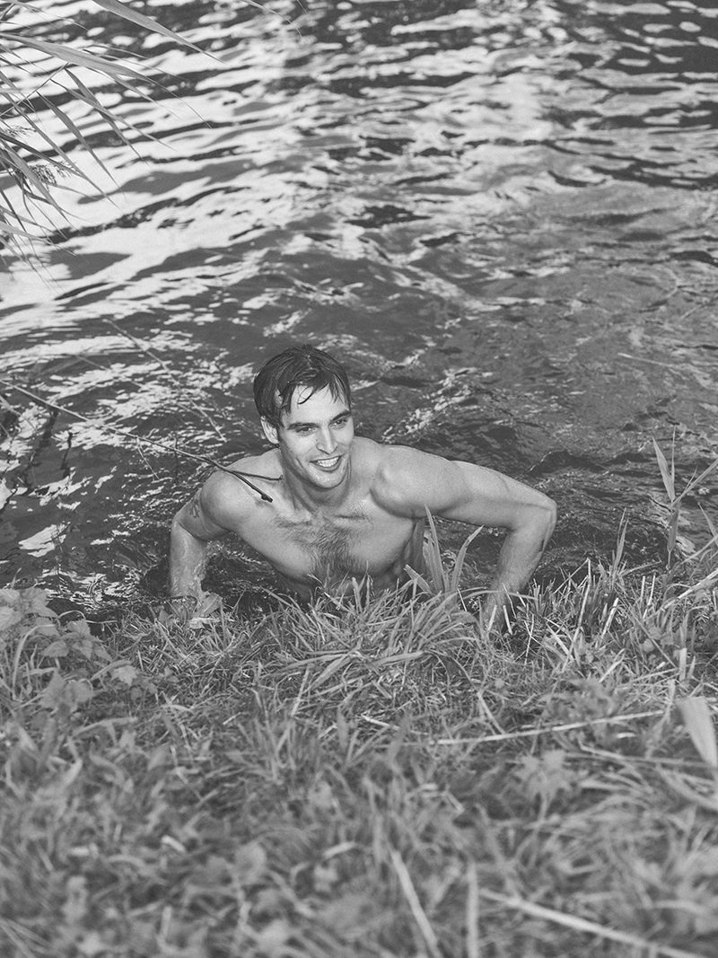 A man with a muscular build is emerging from the water, surrounded by green vegetation in a black and white image, smiling slightly.