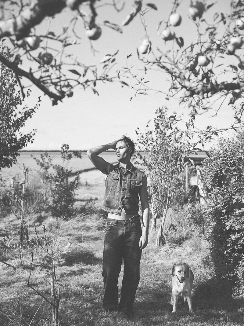 Monochrome portrait of a man in a denim vest standing outdoors with a dog, under an apple tree on a sunny day.